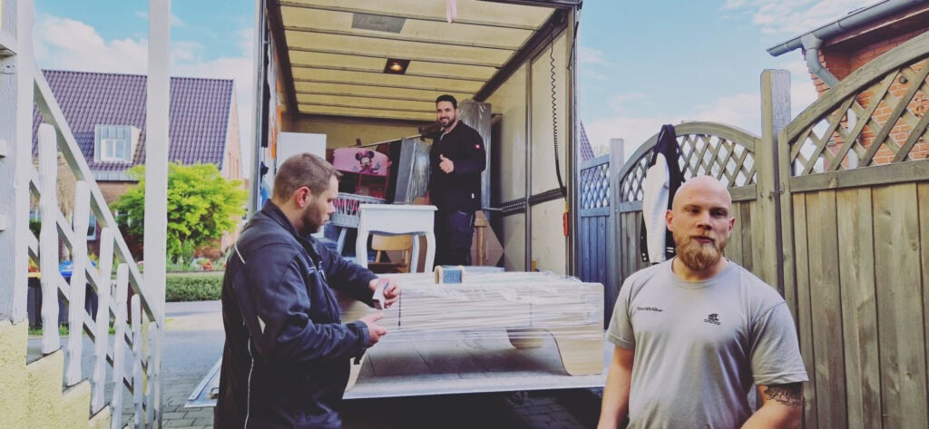 Drei Männer sind bei einem Umzug in Aachen gerade dabei, Möbel zu bewegen. Einer steht mit einem großen Gegenstand auf der Ladefläche eines Lastwagens, ein anderer packt in Plastik eingewickelte Gegenstände in den Lastwagen und der dritte steht mit verschränkten Armen im Vordergrund und blickt aus dem Bild.