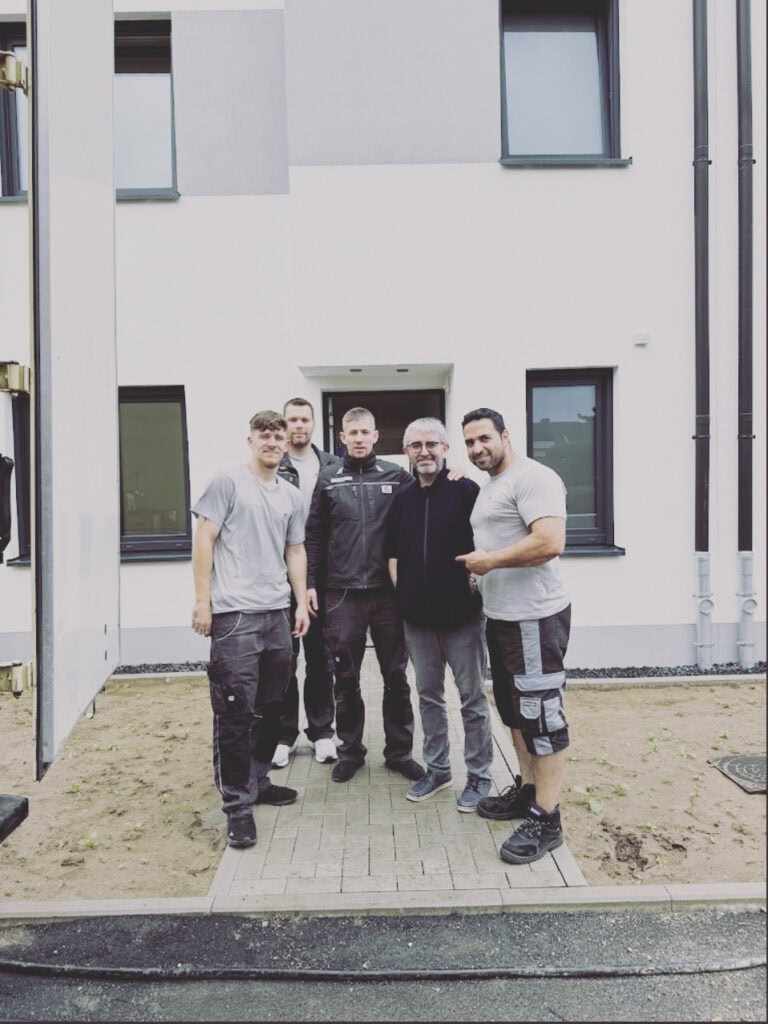 Fünf Männer stehen vor einem modernen weißen Gebäude. Sie tragen legere Arbeitskleidung, manche tragen graue Hemden und Shorts. Die Stimmung wirkt freundlich und entspannt, manche Männer lächeln in die Kamera.